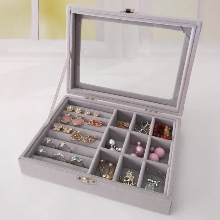 Elegant gray velvet jewelry organizer box with glass lid, displaying rings, earrings, and beads in divided compartments
